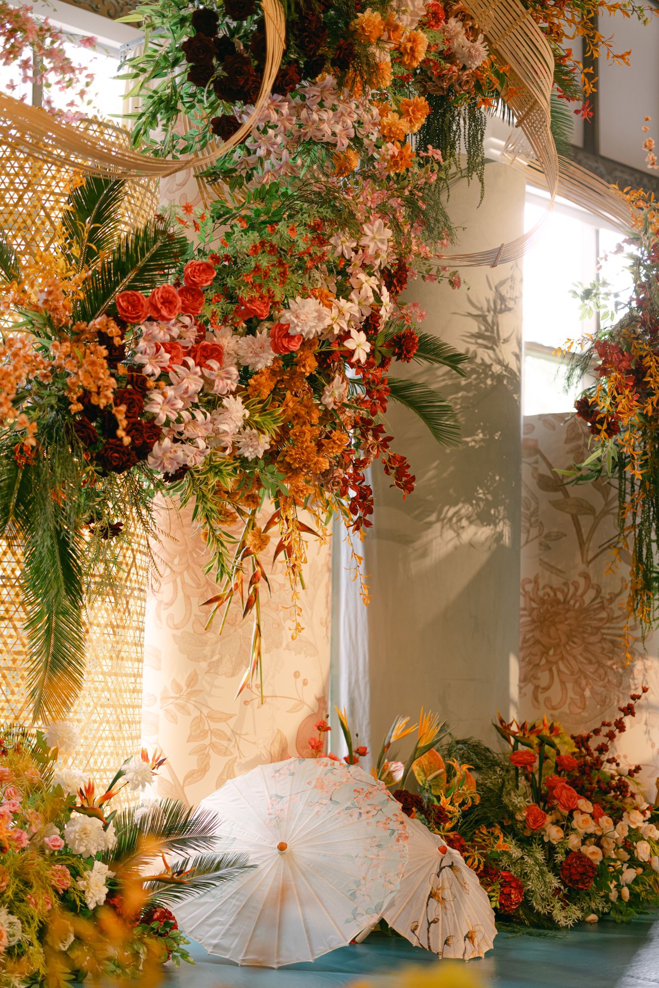 Lush orange, red and pink floral installation hangs above white paper parasols and flower arrangements at a wedding ceremony venue.