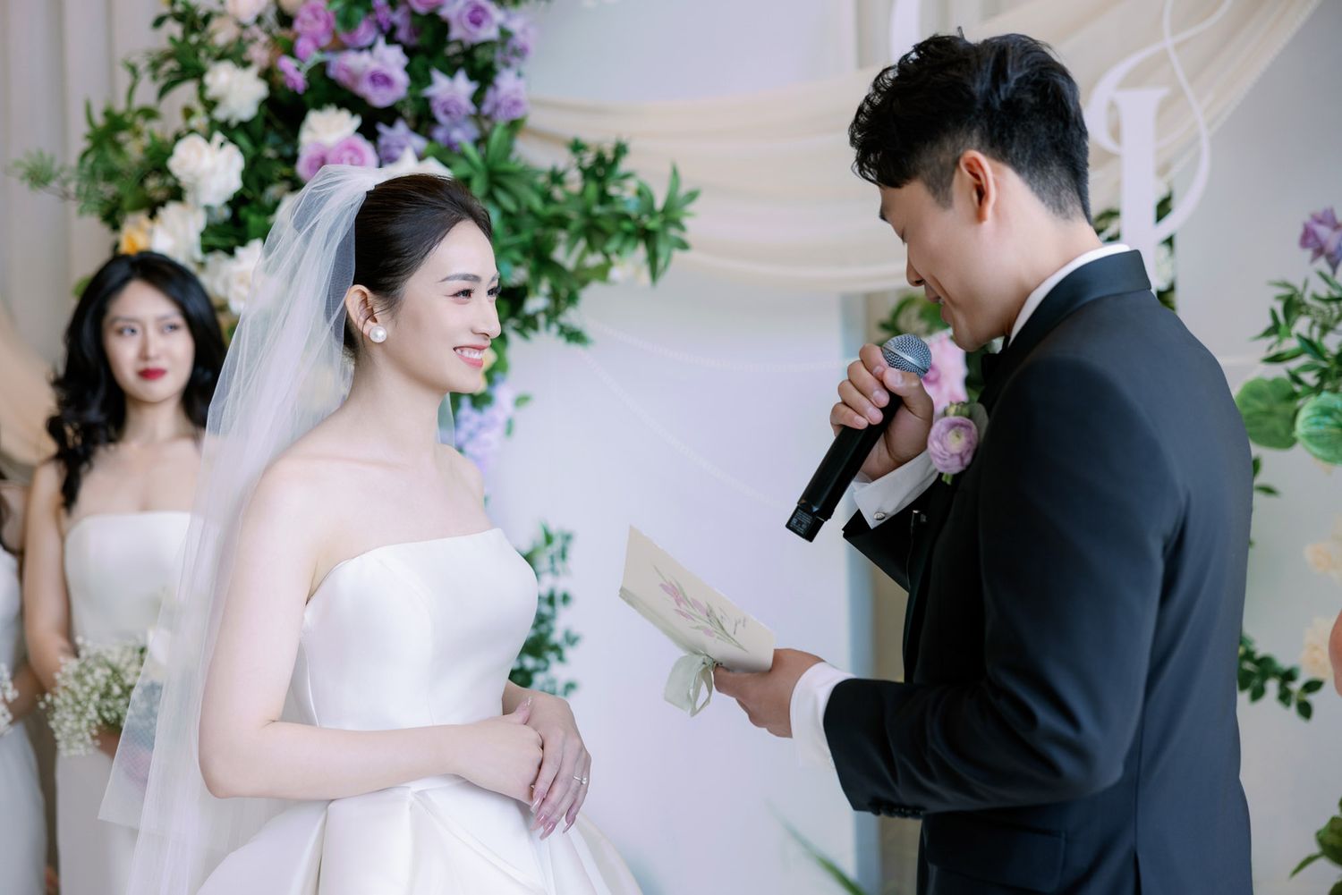 Bride smiles as groom reads heartfelt vows at elegant wedding ceremony with floral backdrop.