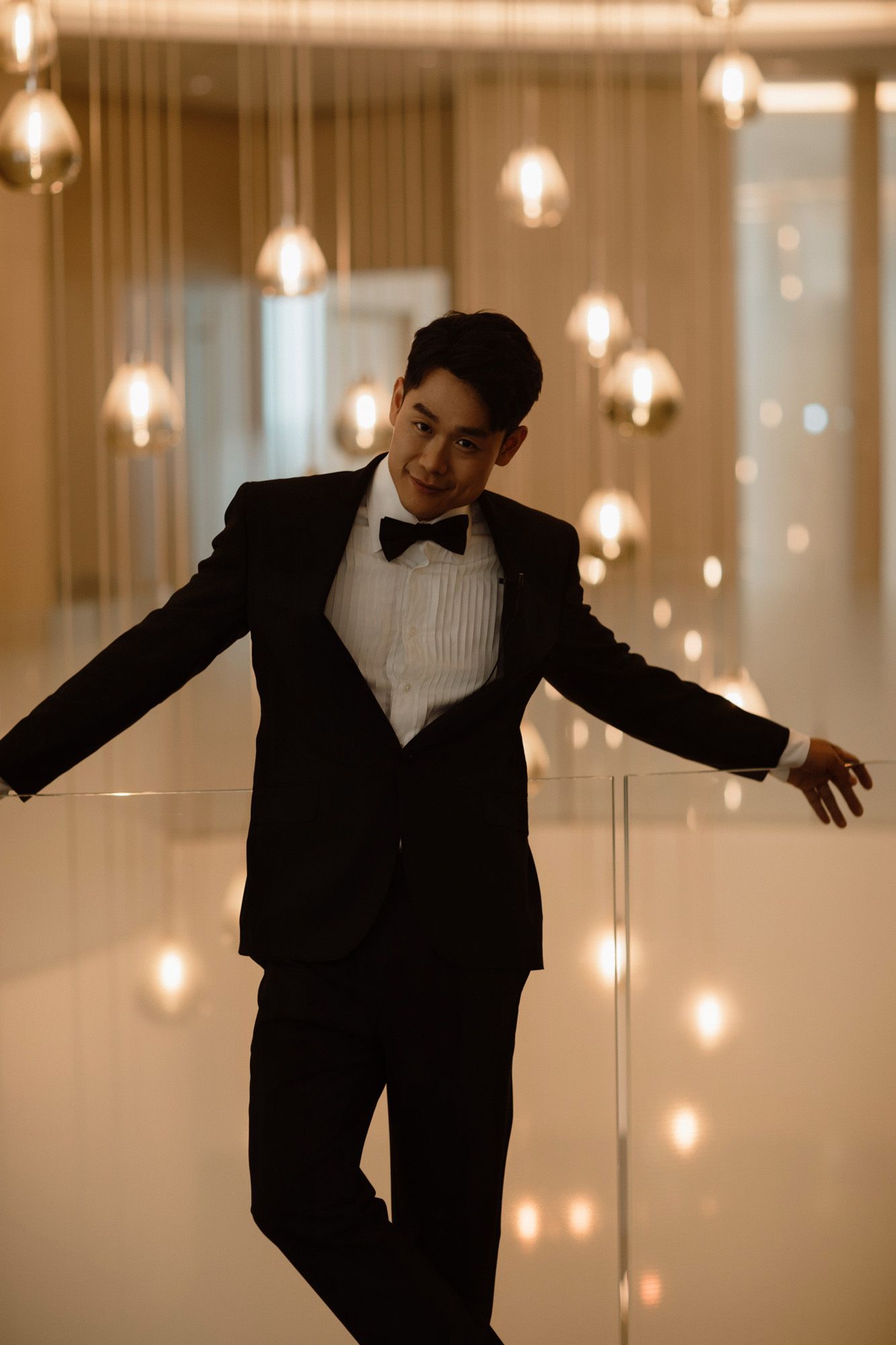 Stylish groom in black tuxedo posing confidently under glowing pendant lights at venue.