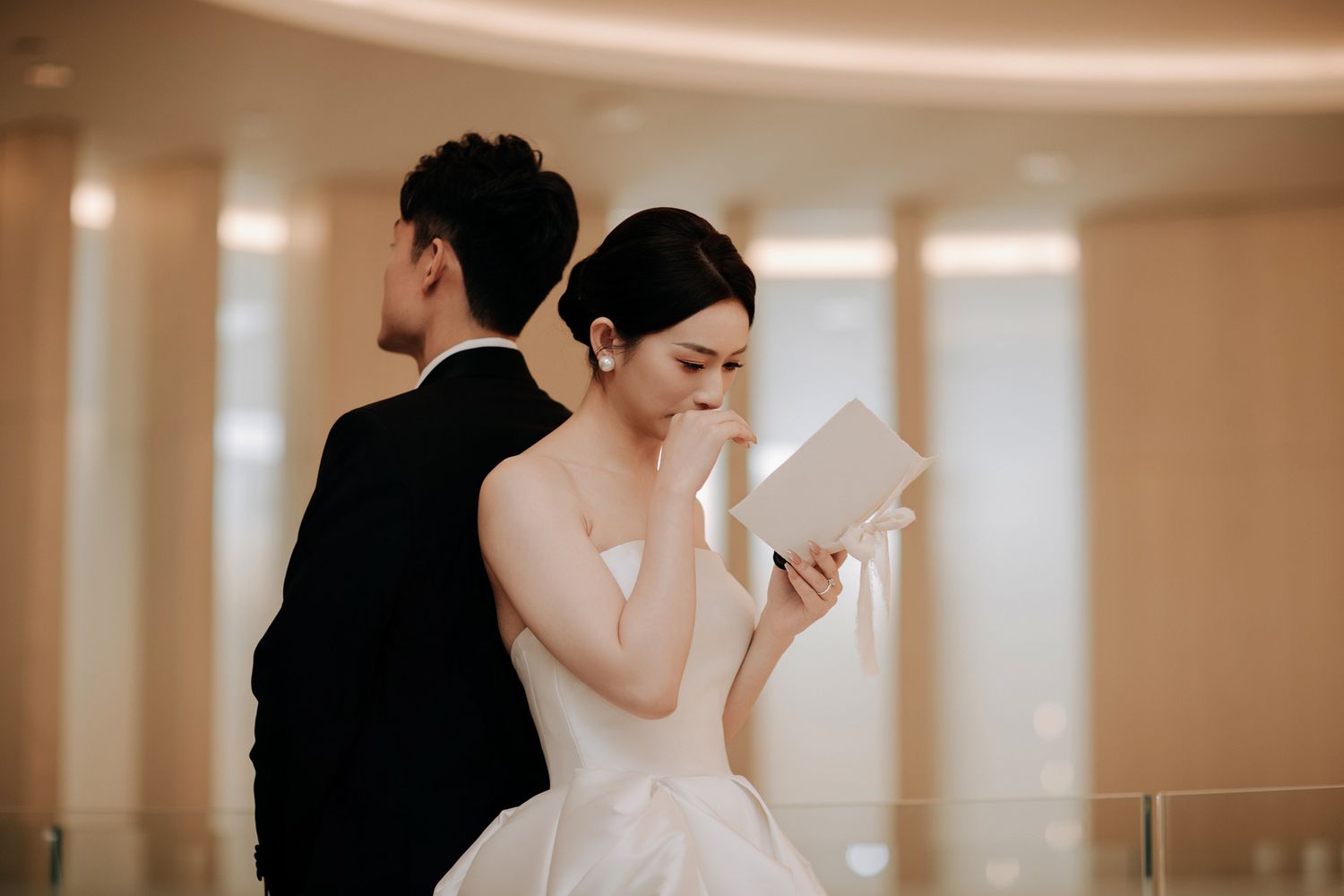 Emotional bride in strapless white gown reads wedding vows letter beside groom in black tuxedo.