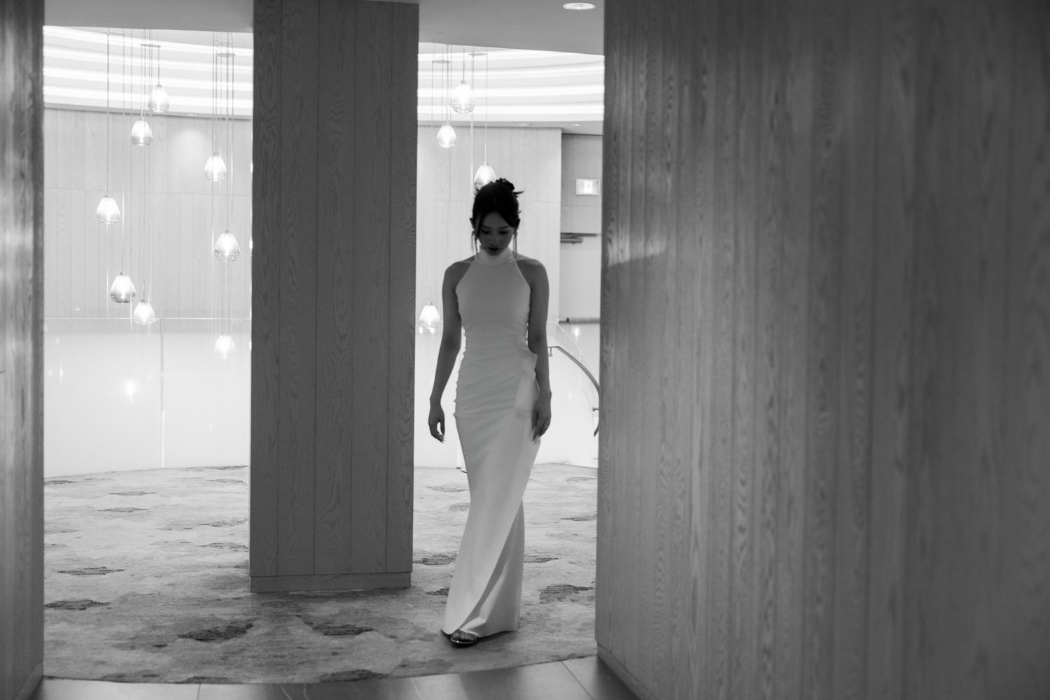 Black and white full length photo of bride in elegant white gown walking through hotel corridor.