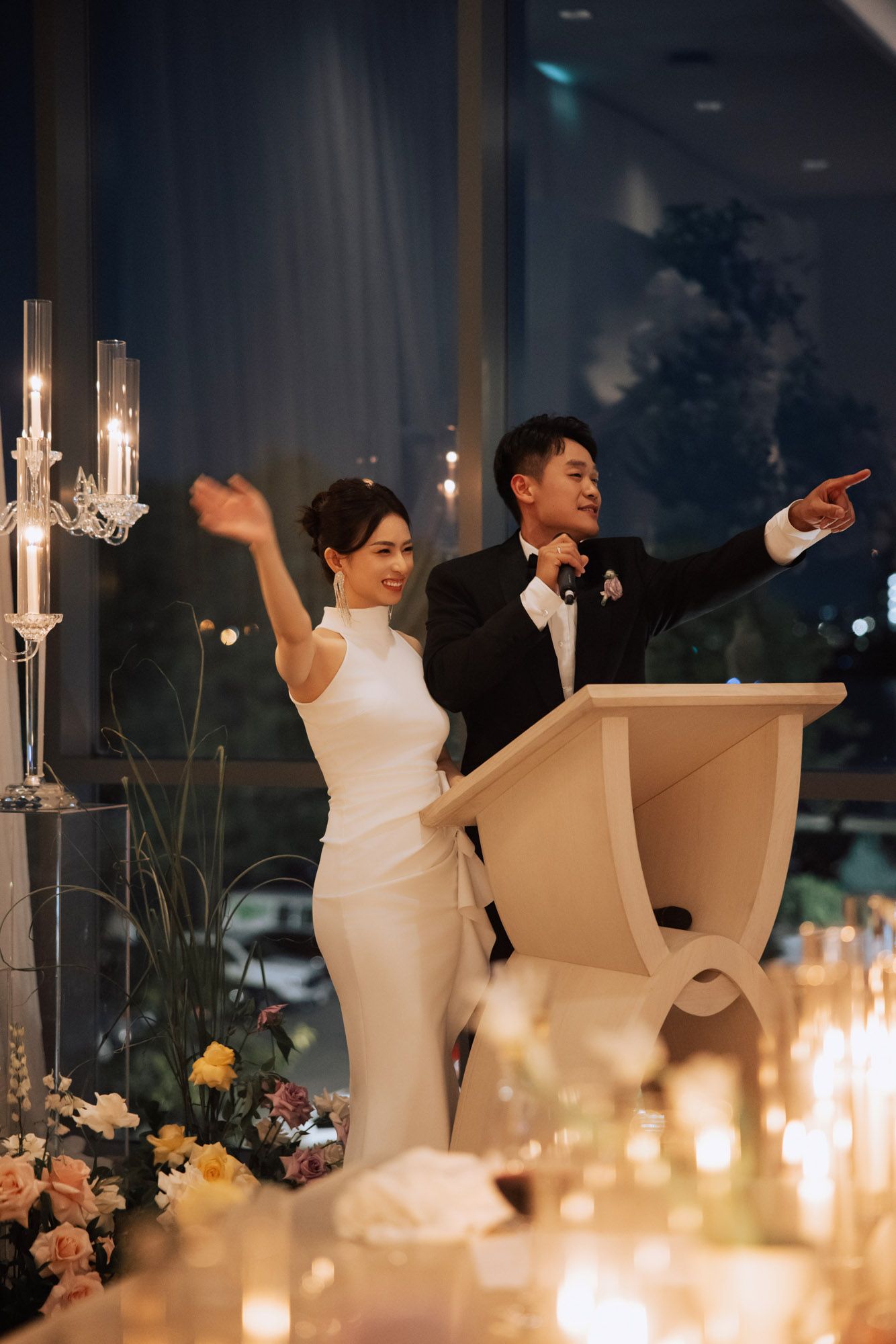 Smiling bride and groom celebrate at wedding reception podium, waving to guests with candles glowing.
