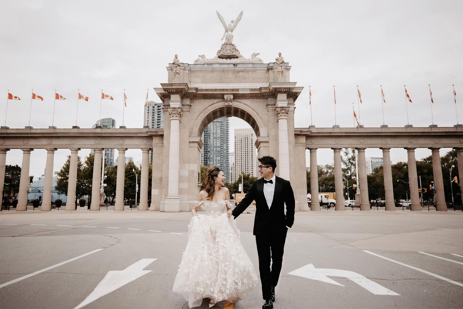 Bride and groom hold hands before ornate stone arch at Toronto's CNE grounds on their wedding day.