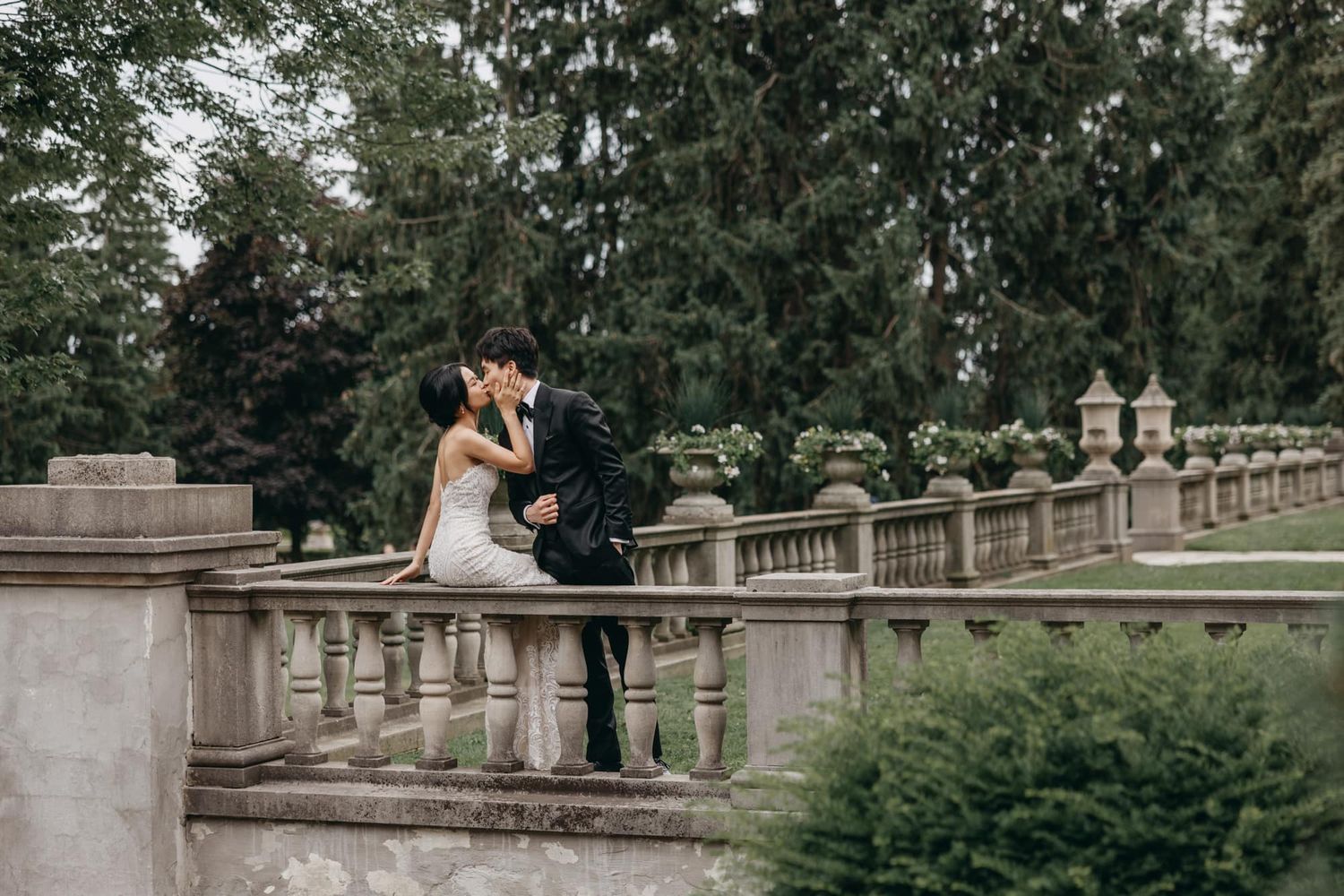 A bride and groom share a romantic kiss on a stone balustrade in a lush garden during their wedding day.