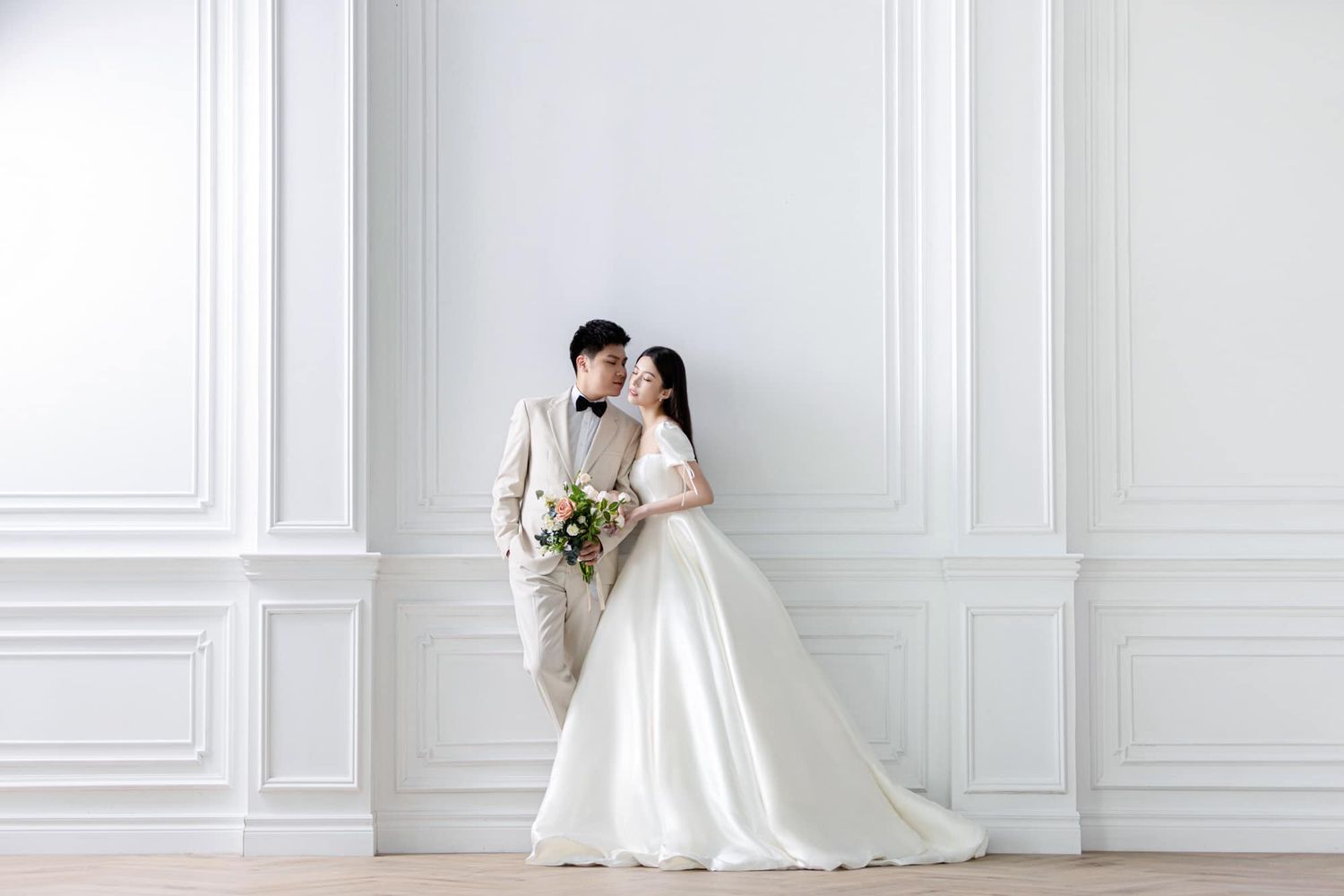 A couple in wedding attire poses elegantly against a white paneled wall, the bride in a flowing gown holding a bouquet.