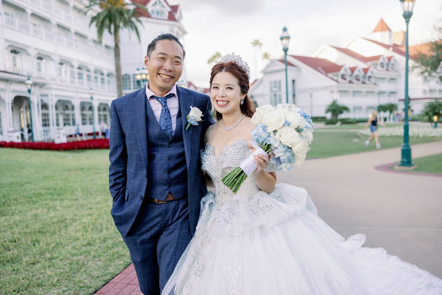Couple poses in wedding attire outside Disney's Grand Floridian Resort, bride holding white bouquet.