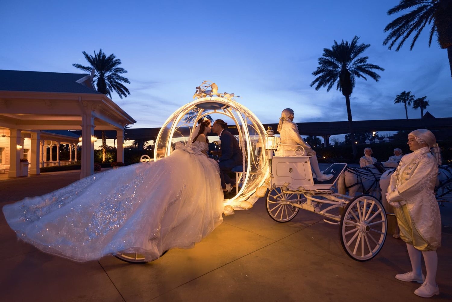 Bride in white gown boarding a glowing Cinderella carriage at dusk, with palm trees and elegant venue in background.