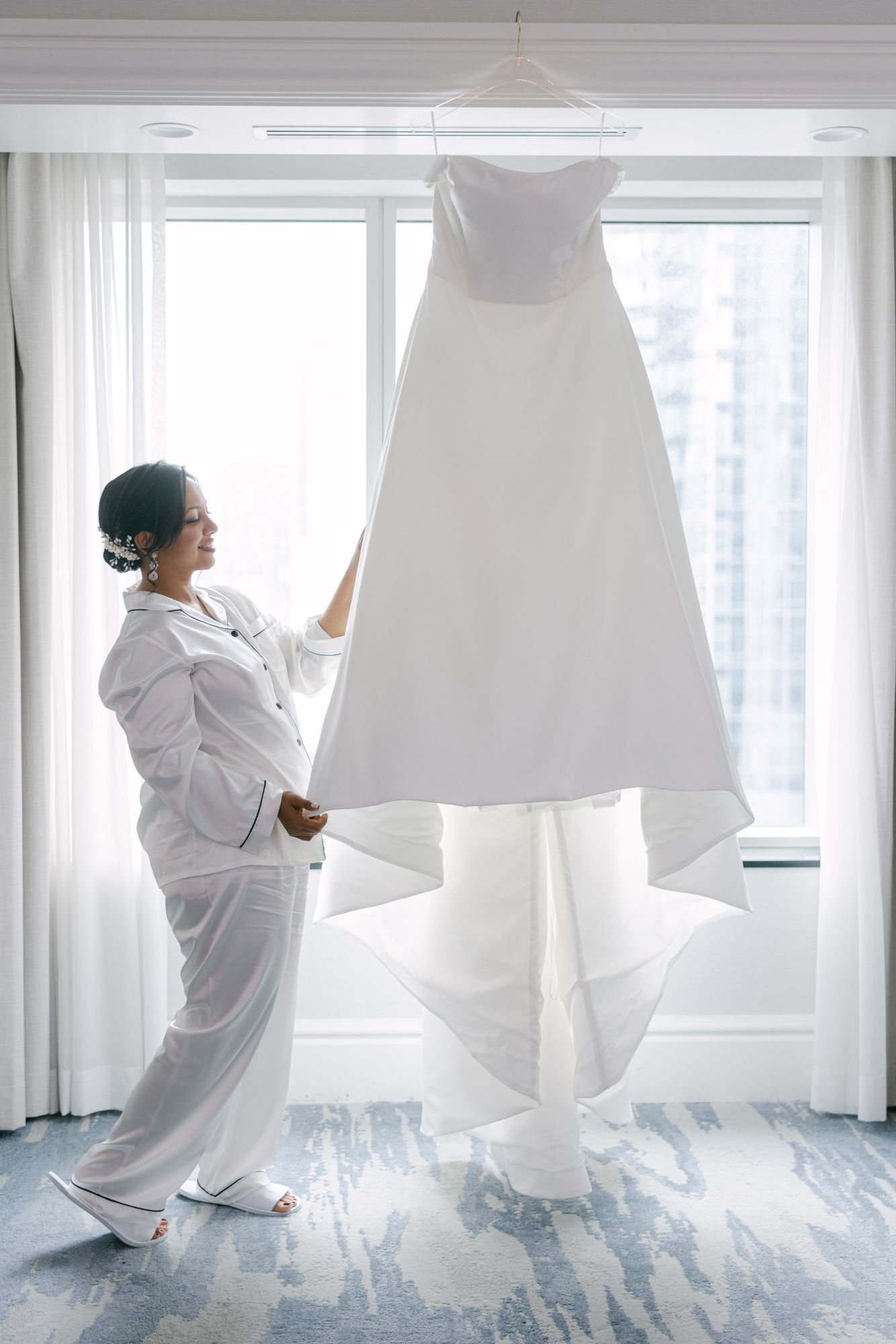 Bride in white pajamas gazes at her strapless wedding gown hanging from a window in a bright hotel room before the ceremony.