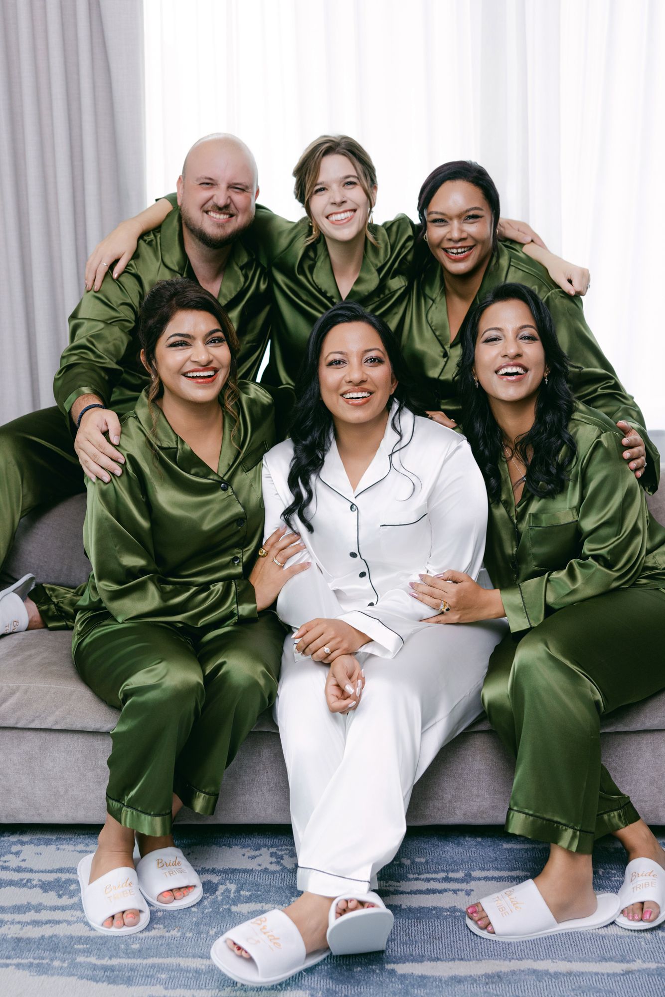 Bride in white satin pajamas sits smiling on a gray sofa surrounded by five friends in matching green silk pajamas and white slippers.