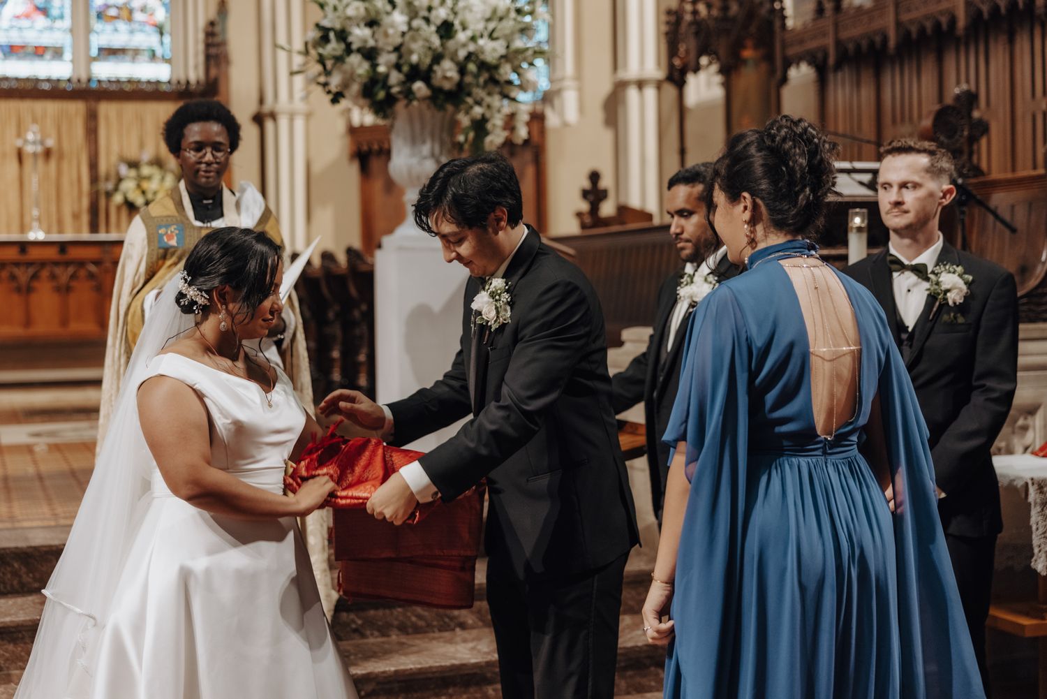 Bride and groom exchange a red cloth during a church wedding ceremony as guests in blue and black look on.