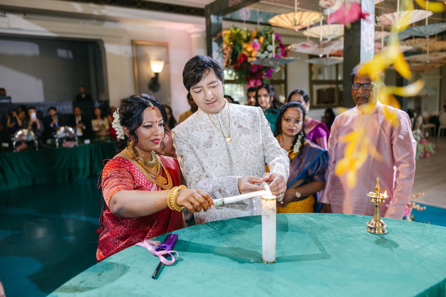 A bride in a red and orange saree lights a candle with a groom in an ivory sherwani during an Indian wedding ritual as guests look on.