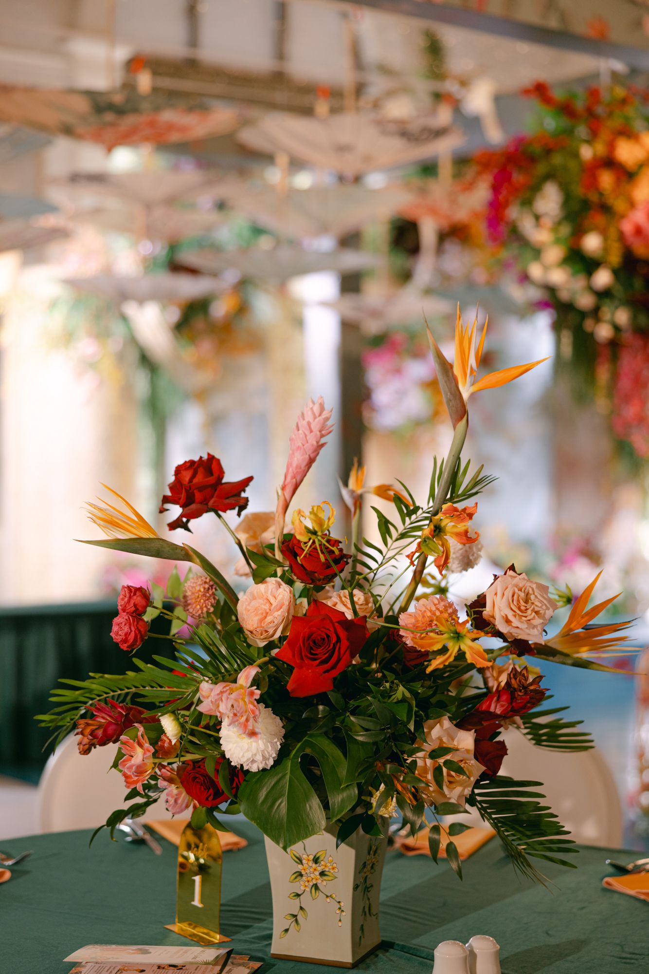 Wedding reception centerpiece with red roses, orange bird of paradise and greenery in a vase on a green tablecloth.