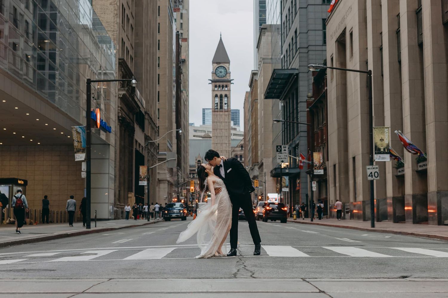 Bride and groom sharing a romantic dip kiss on a city crosswalk with a historic clock tower backdrop.