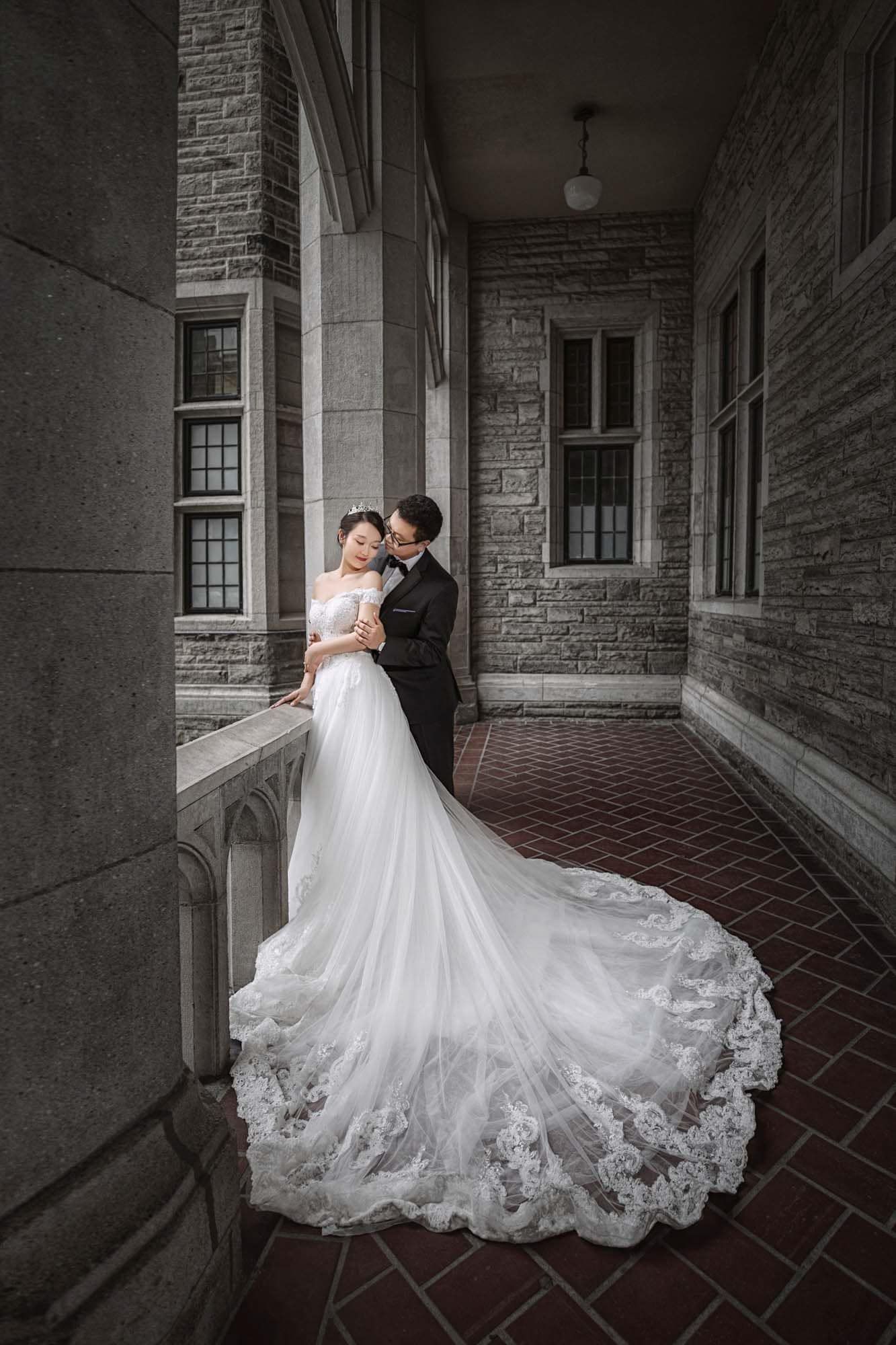 Bride in off-shoulder gown with long lace train embracing groom on castle balcony corridor.
