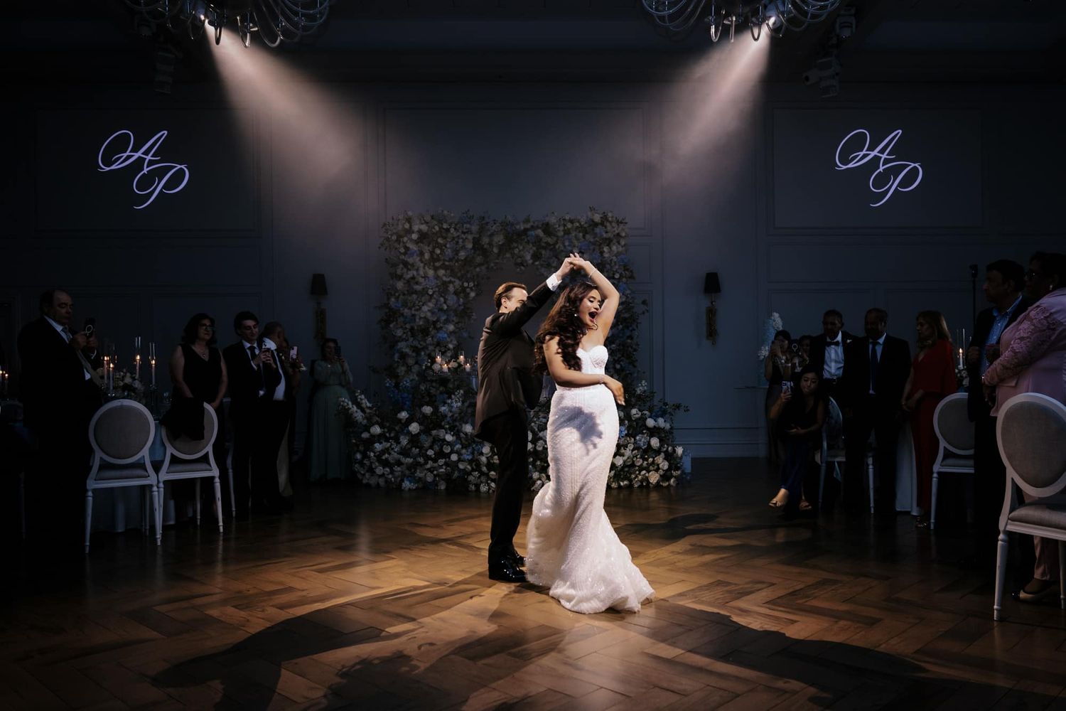 Bride and groom dance elegantly under monogram lighting with blue floral arrangements at wedding.