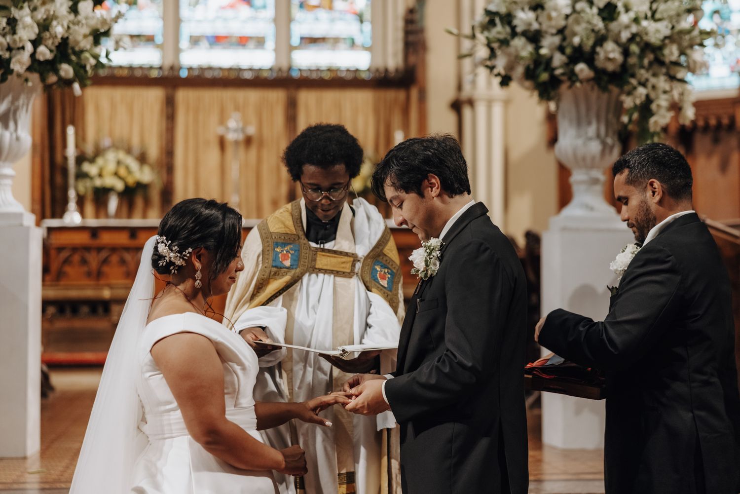 A bride in a white veil and groom exchange rings during a church wedding ceremony led by a priest in ceremonial vestments.