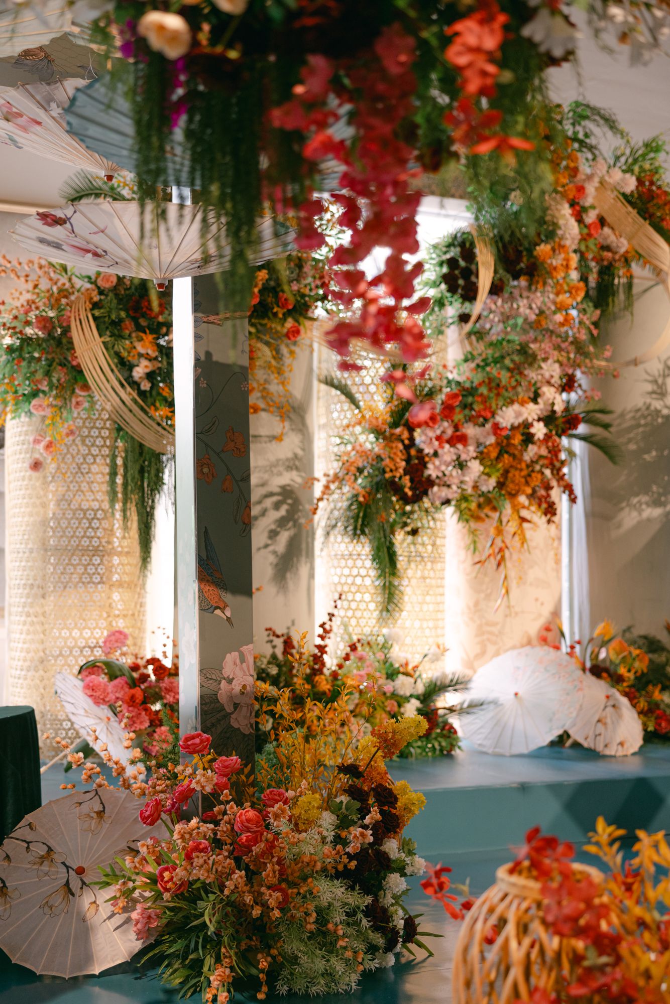 Wedding stage decorated with hanging red, orange and pink floral arrangements, white paper parasols and lattice backdrops.
