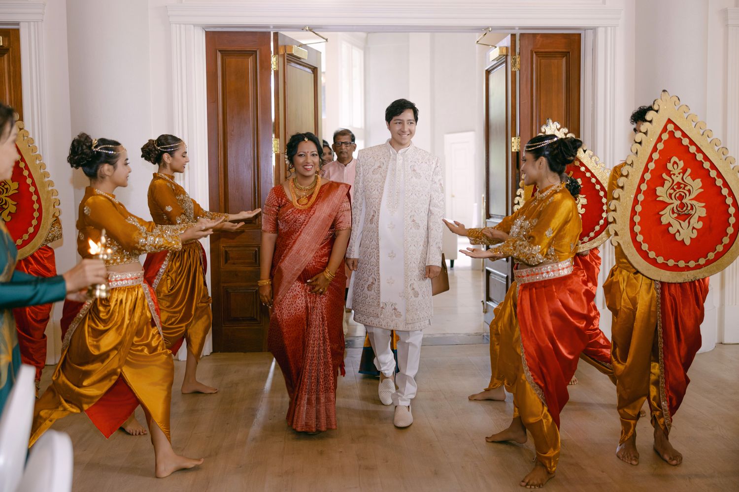 Traditional dancers in gold and orange costumes greet a woman in a red sari and a man in a white tunic at a grand entrance.