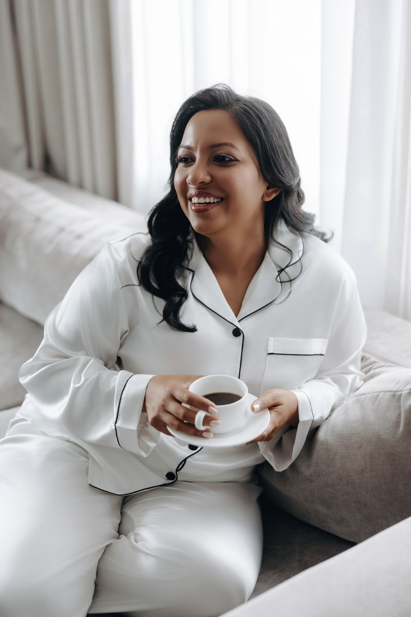 Smiling woman with long dark wavy hair in white satin pajamas sits on a gray sofa holding a cup of coffee by sheer curtains.
