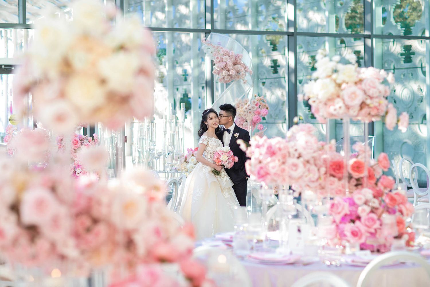 Bride and groom stand surrounded by lush pink floral arrangements and elegant table settings in a dreamy wedding venue.