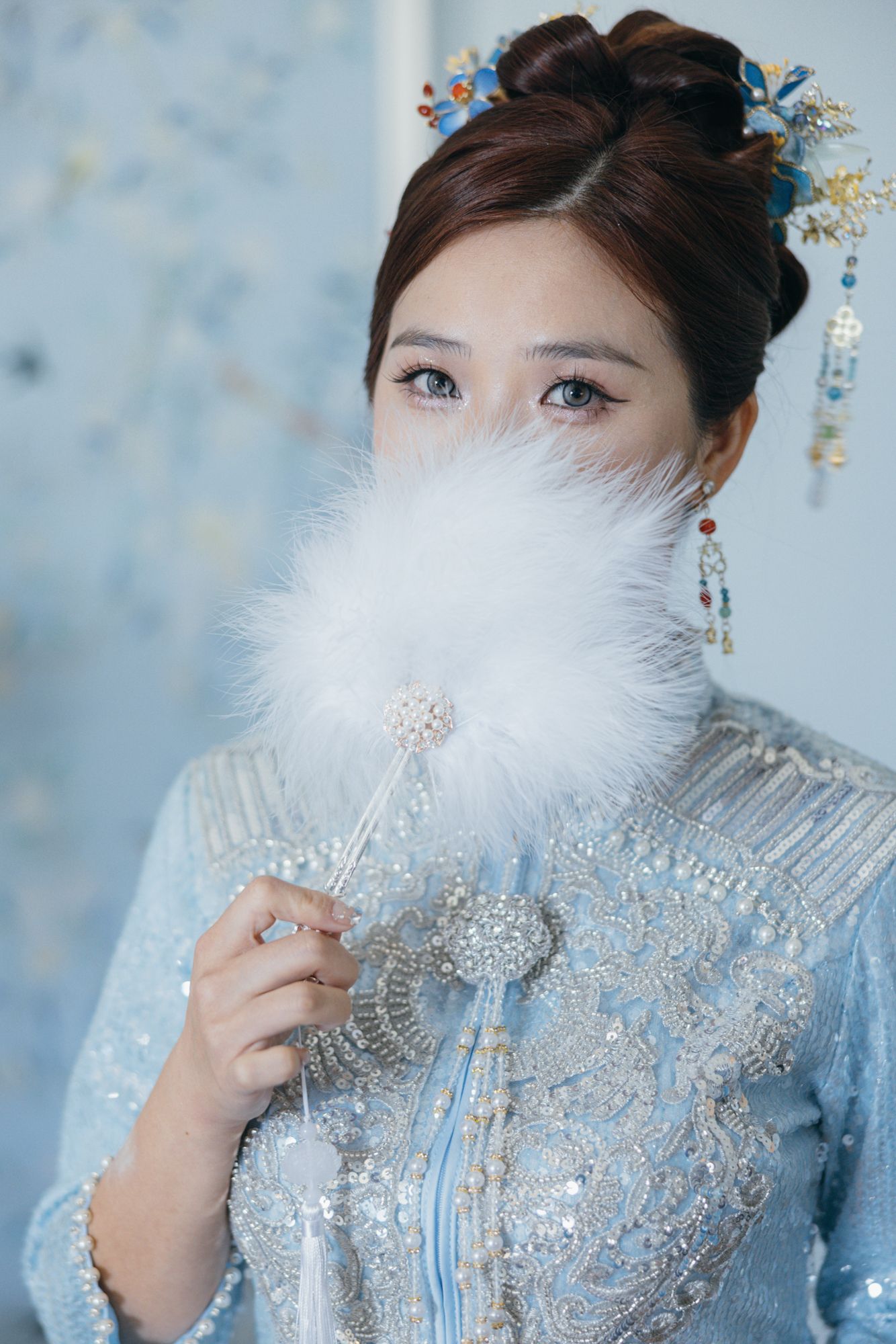 Bride in light blue Chinese wedding dress holds white feather fan covering her face, adorned with floral hair accessories.