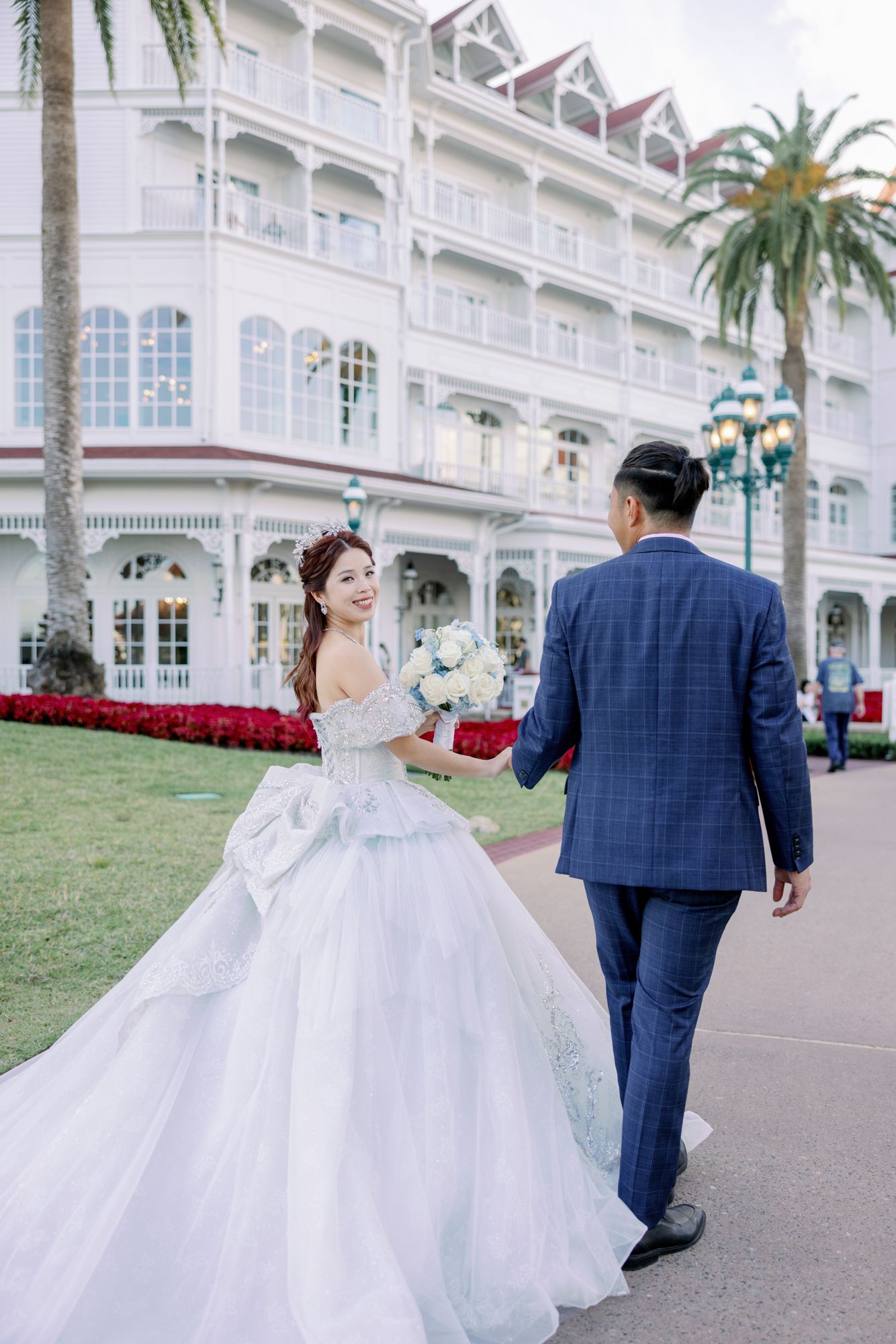 Bride in a stunning ball gown and groom in a blue suit walk hand-in-hand outside a grand white Victorian hotel.