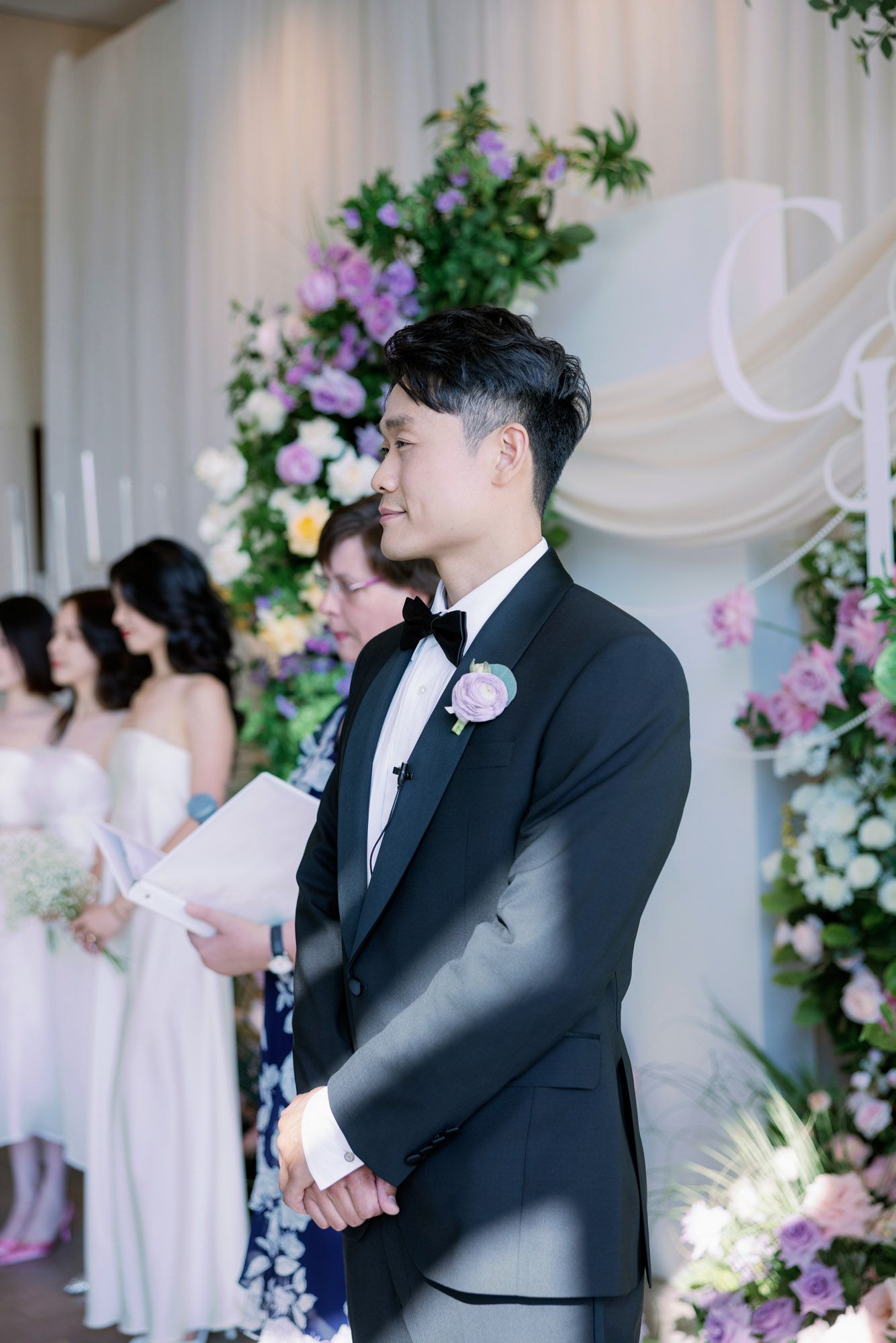 Groom in black tuxedo smiling at altar with bridesmaids and floral arch at elegant wedding ceremony.