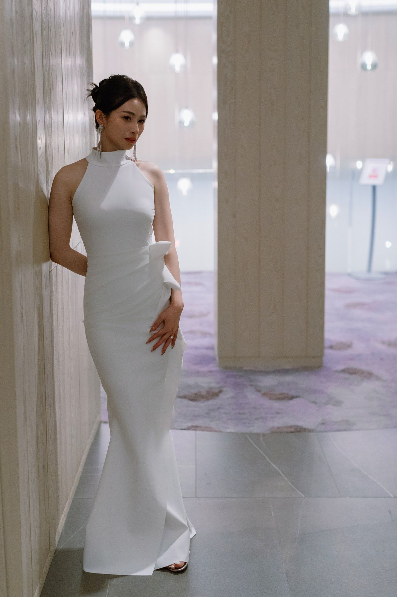 Elegant bride in sleek white halter neck gown posing gracefully against marble column in hotel.