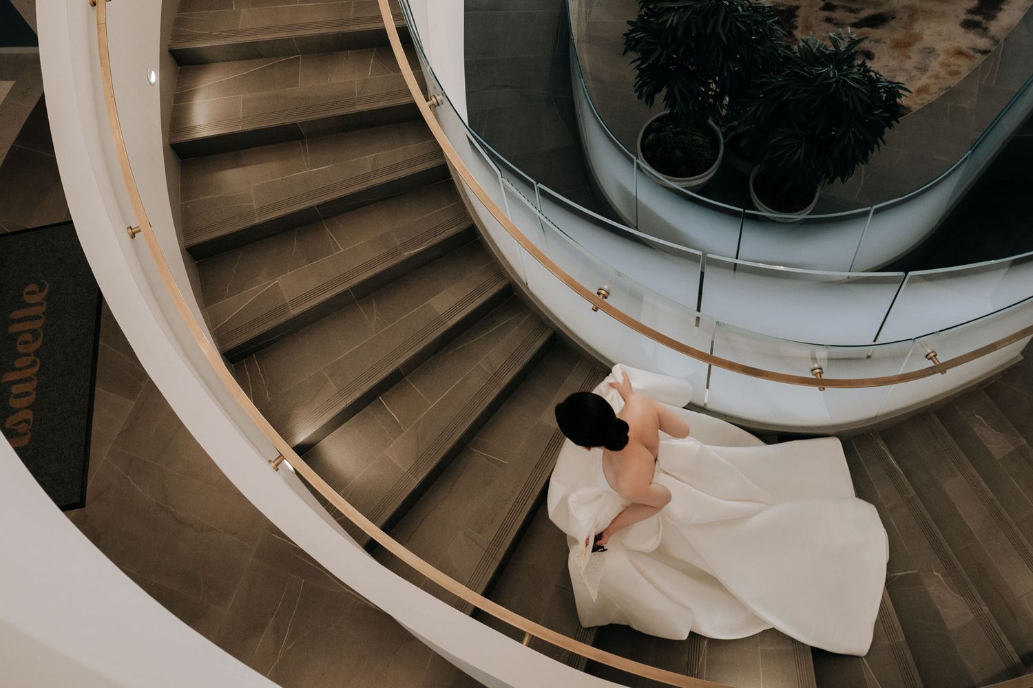 Top-down view of bride in flowing white wedding dress sitting on curved spiral staircase.