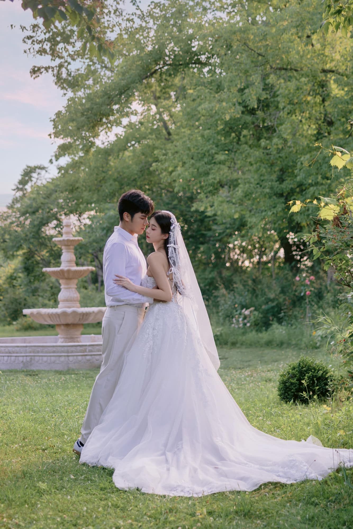 Bride and groom embrace in lush garden with stone fountain, bride wearing elegant white gown with veil and long train.