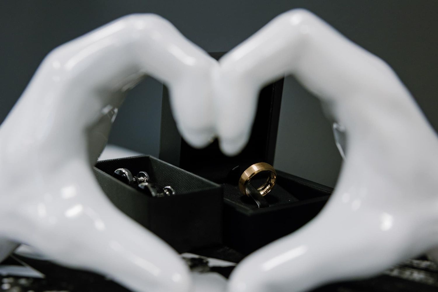 Ceramic hands forming a heart shape around a gold wedding ring displayed in a black jewelry box.