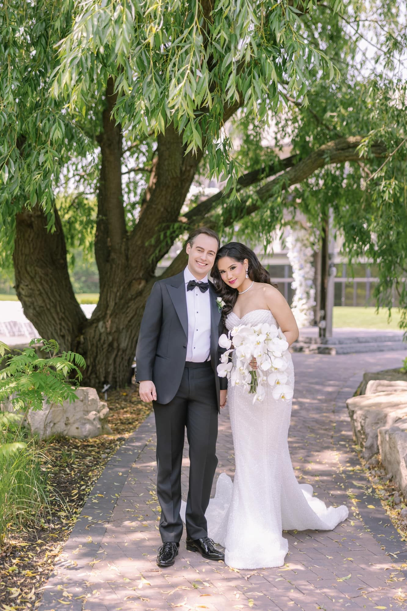 Bride and groom posing under weeping willow tree on brick path at elegant outdoor wedding.