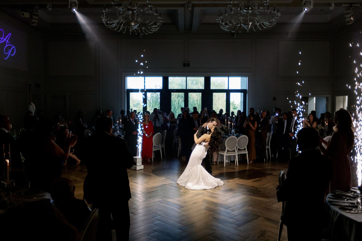Bride and groom share first dance surrounded by guests in elegant ballroom with sparkling lights.