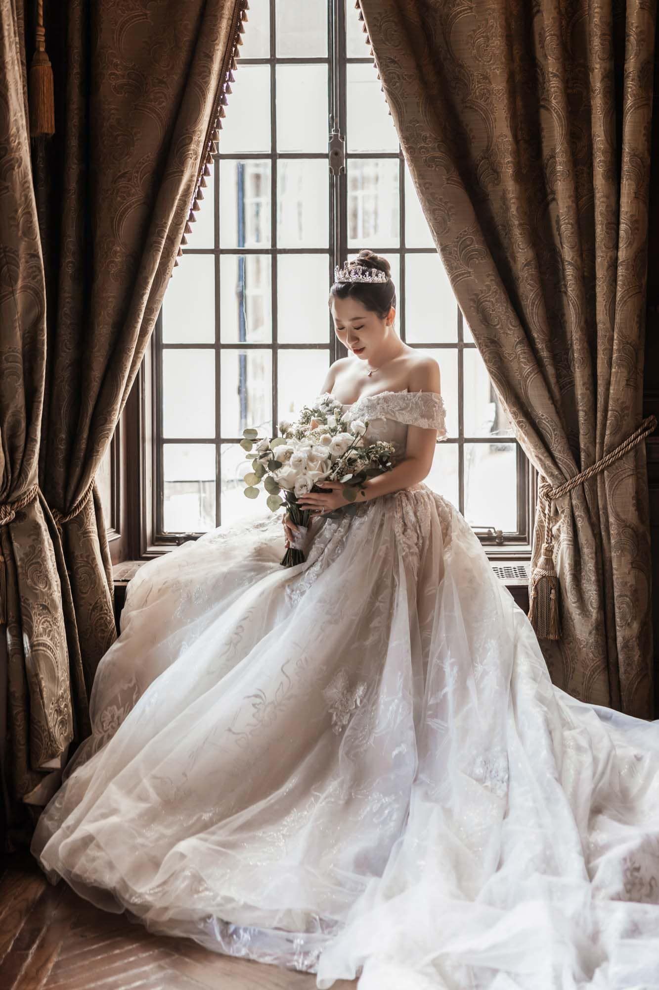 Beautiful Asian bride in ornate off-shoulder gown holding bouquet by a large vintage window.