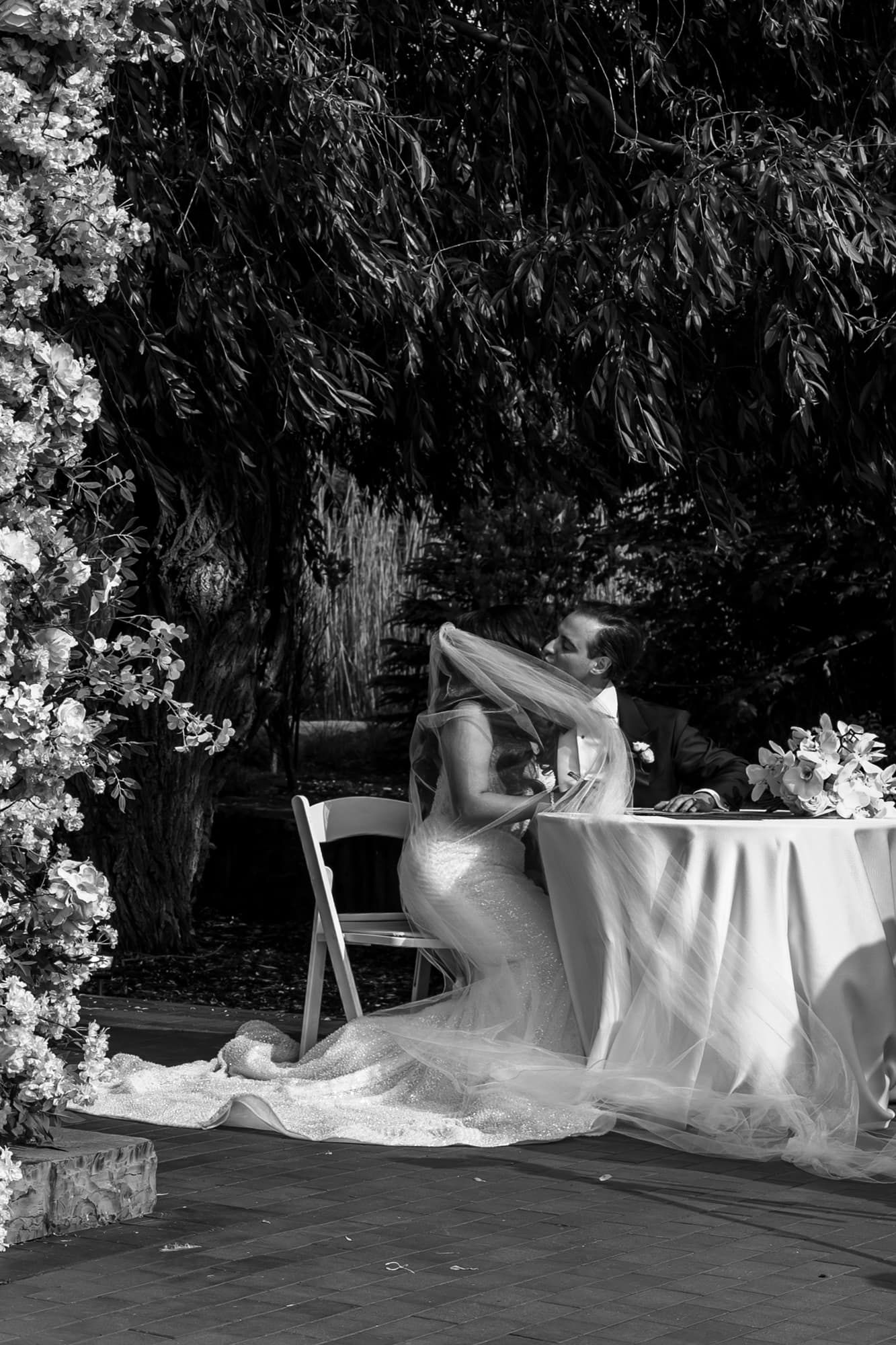 Black and white photo of bride and groom signing wedding documents outdoors.