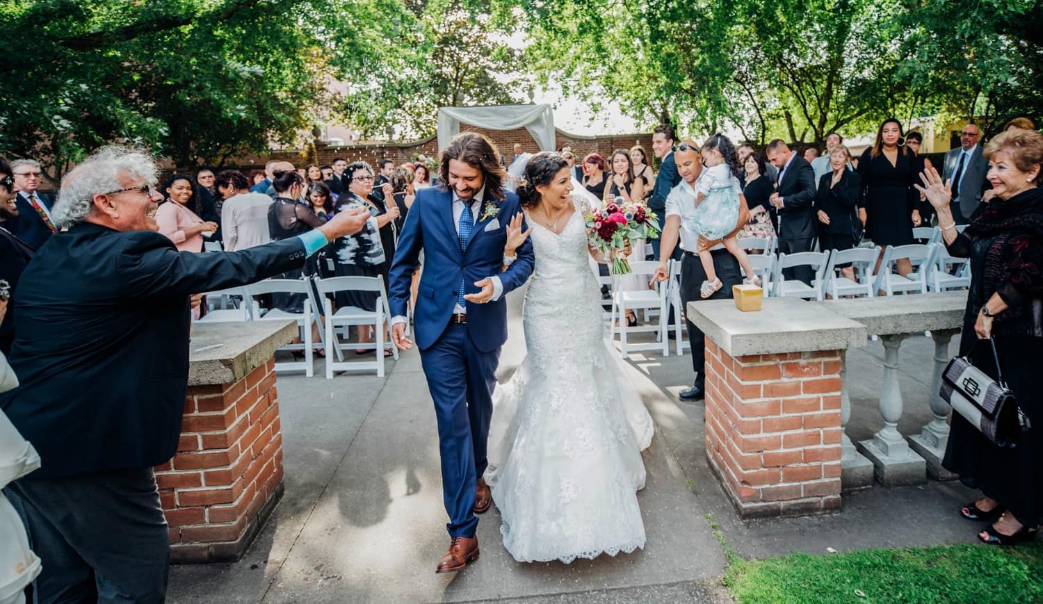 Newlyweds walk joyfully through an outdoor garden ceremony with guests cheering around them.