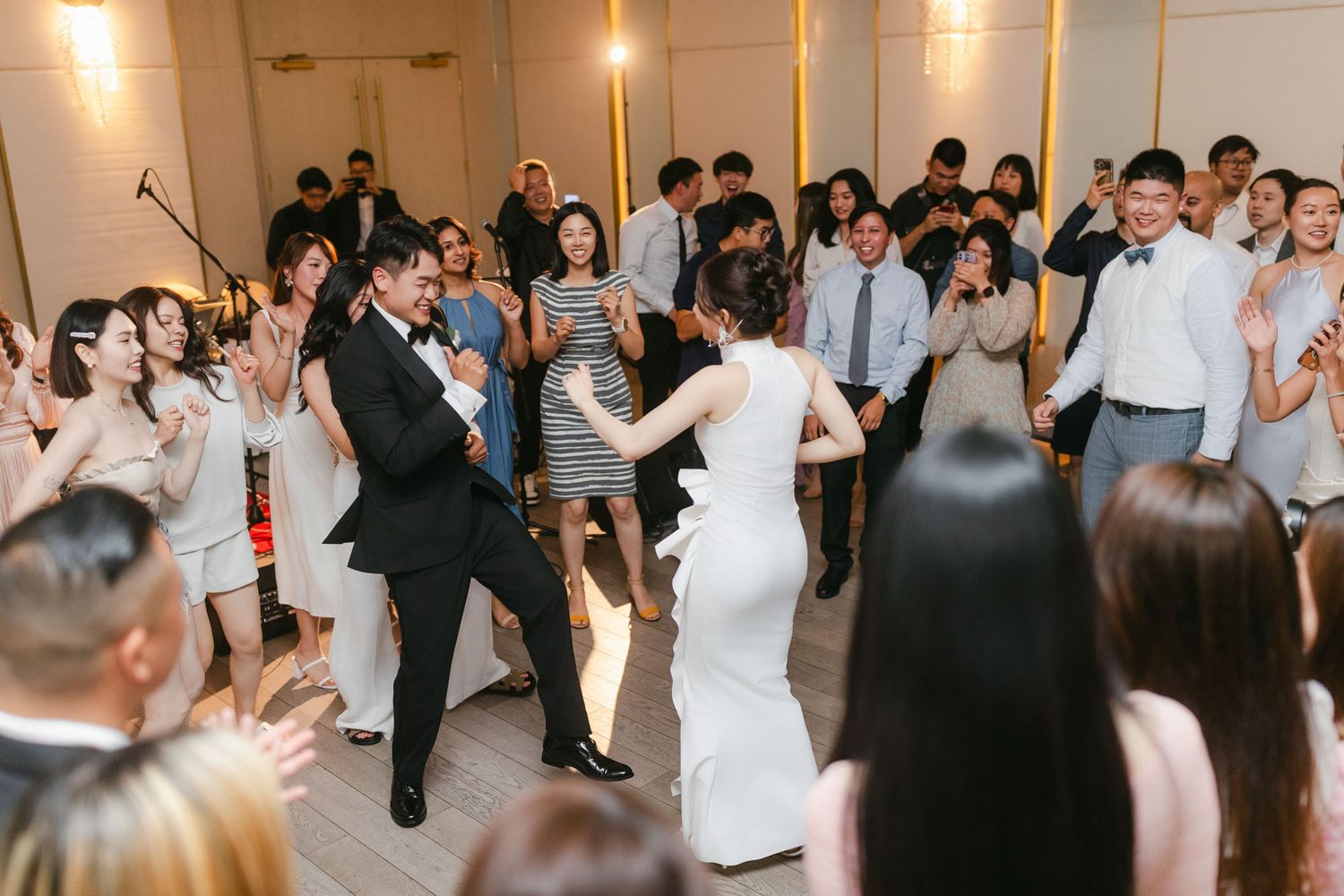 Bride and groom dance joyfully surrounded by cheering wedding guests in elegant venue.