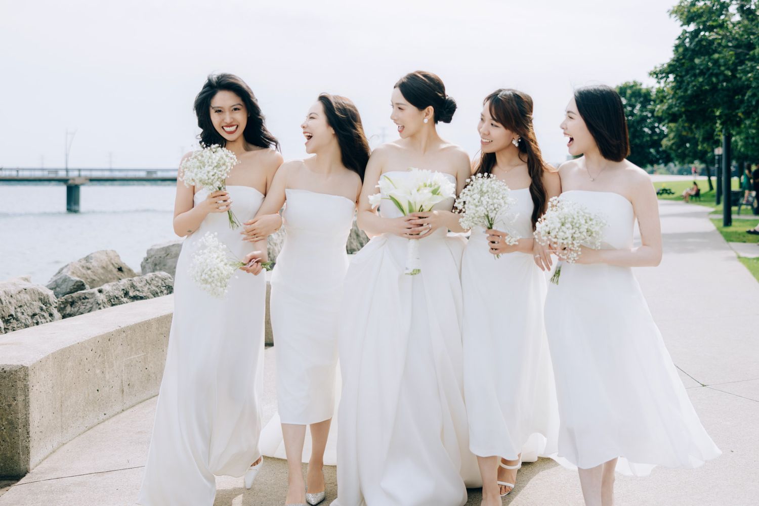 Bride and four bridesmaids in matching white dresses laugh together outdoors near waterfront.