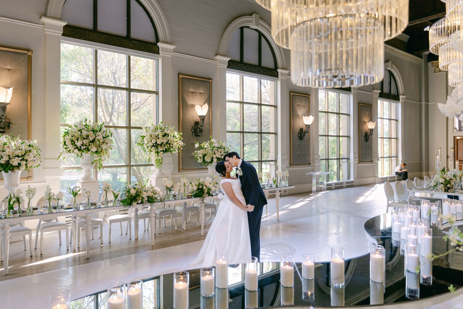 Bride and groom embrace in an elegant ballroom with crystal chandeliers, tall windows, and floral-adorned reception tables.