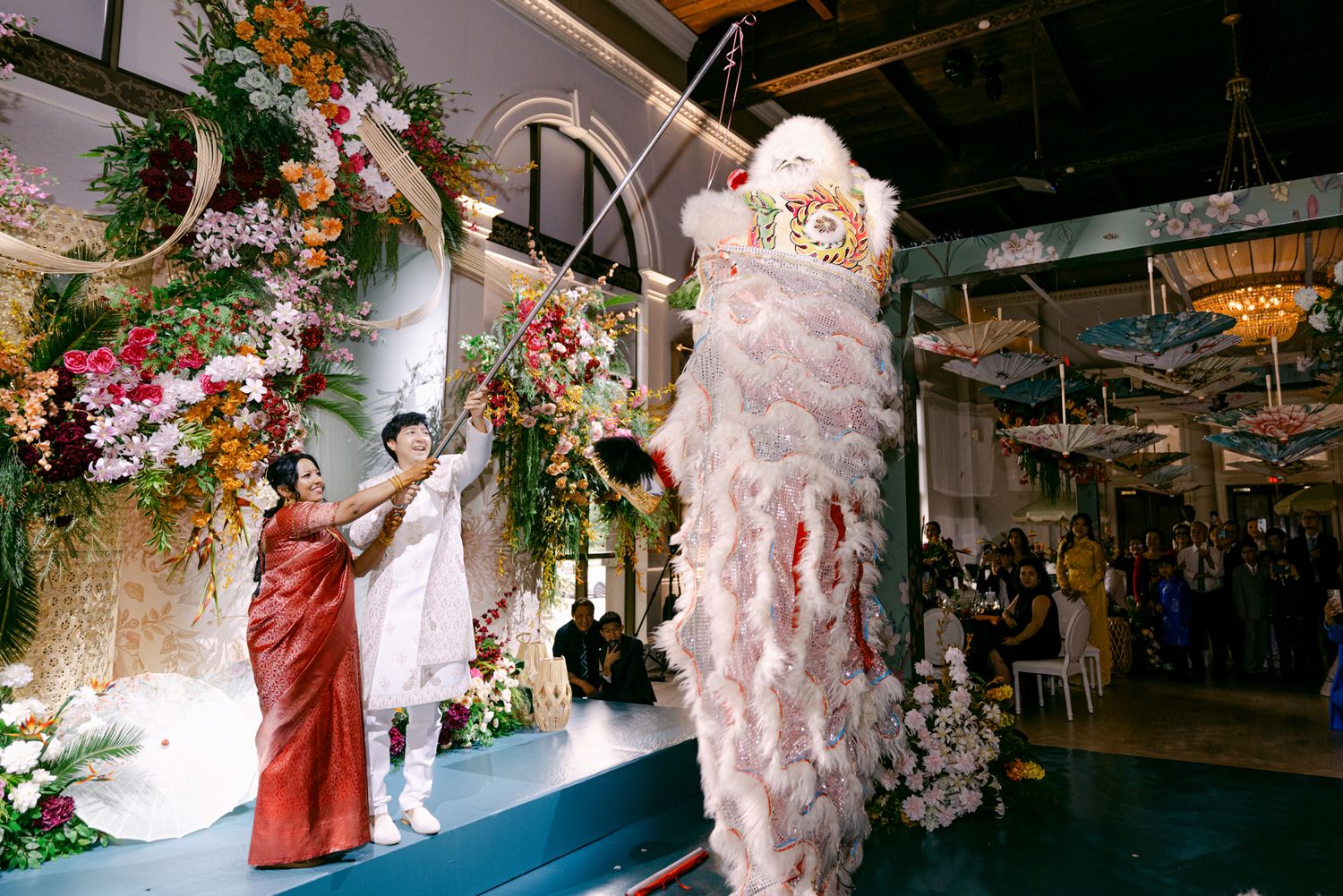 Performers animate a white and red lion dance costume at a wedding while a woman in red reaches toward it beside a floral display.