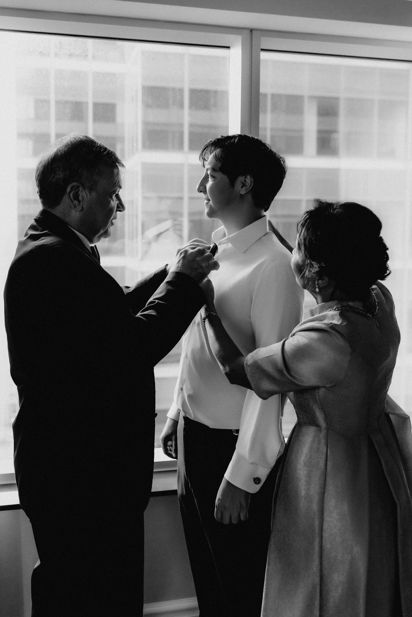 Black and white photo of a groom getting ready by a window as his parents adjust his shirt and boutonniere.