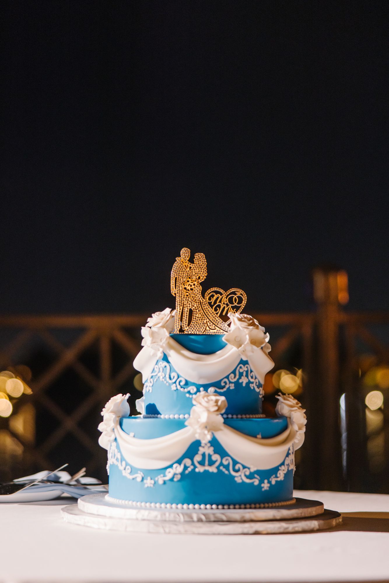 Elegant two-tier blue wedding cake with white lace details and gold couple topper on dark background.