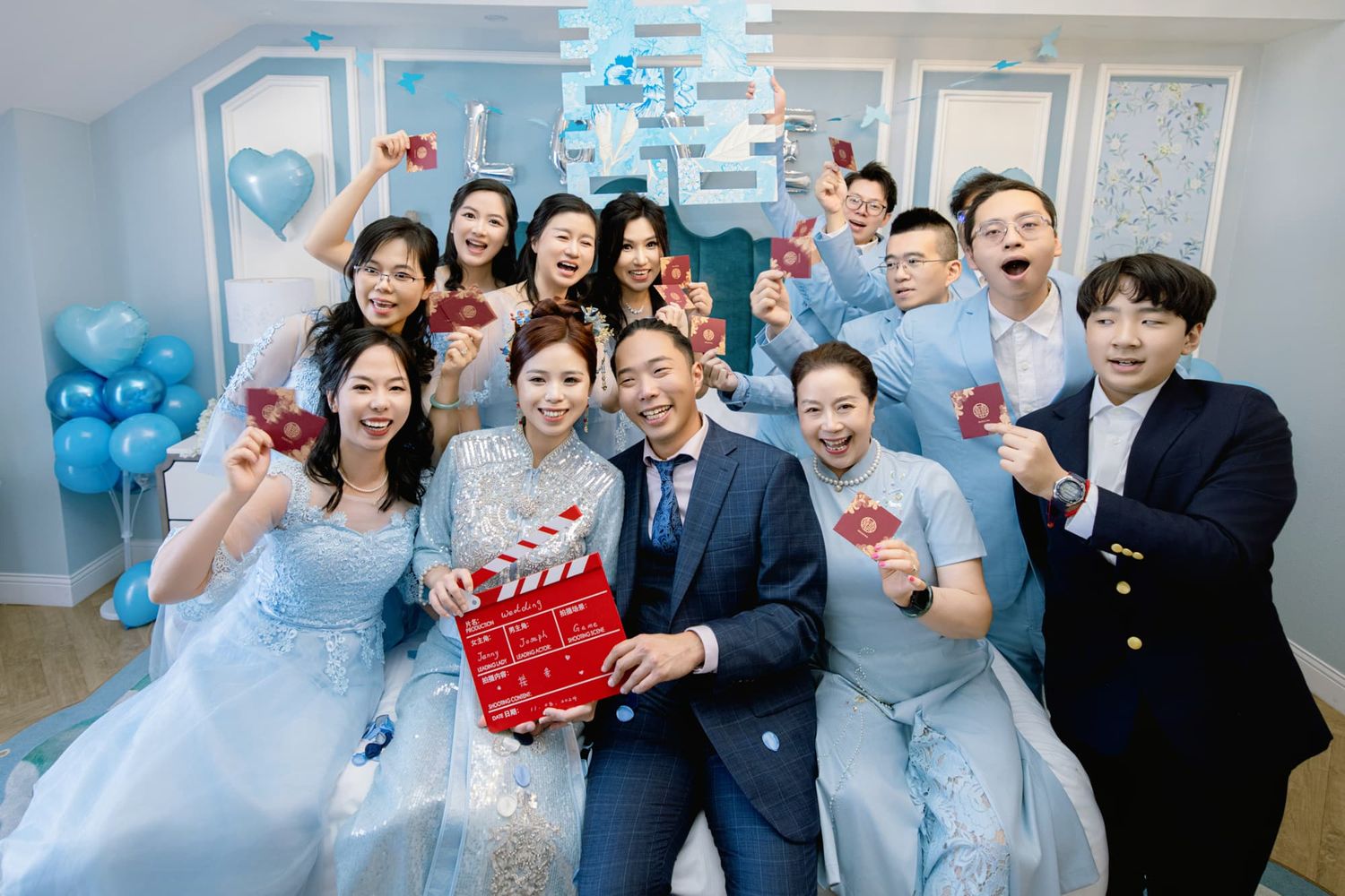 Wedding party group photo with couple holding a red clapperboard in a blue-themed decorated room.