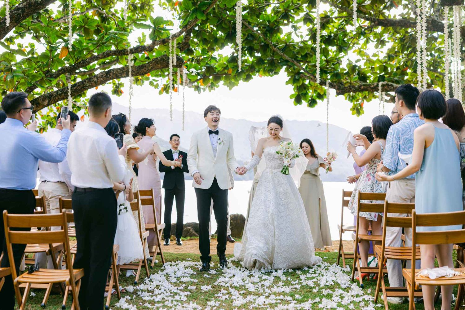Bride and groom celebrate their outdoor ceremony exit as guests toss petals beneath a lush tree.