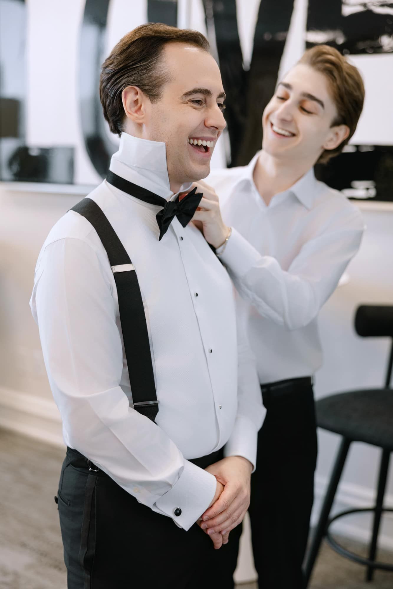 Best man helping laughing groom adjust black bow tie while getting ready for wedding.