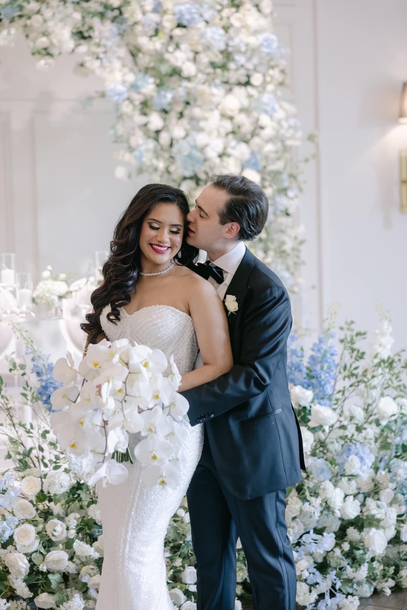 Groom tenderly kisses bride holding white orchid bouquet before lush floral arrangement.