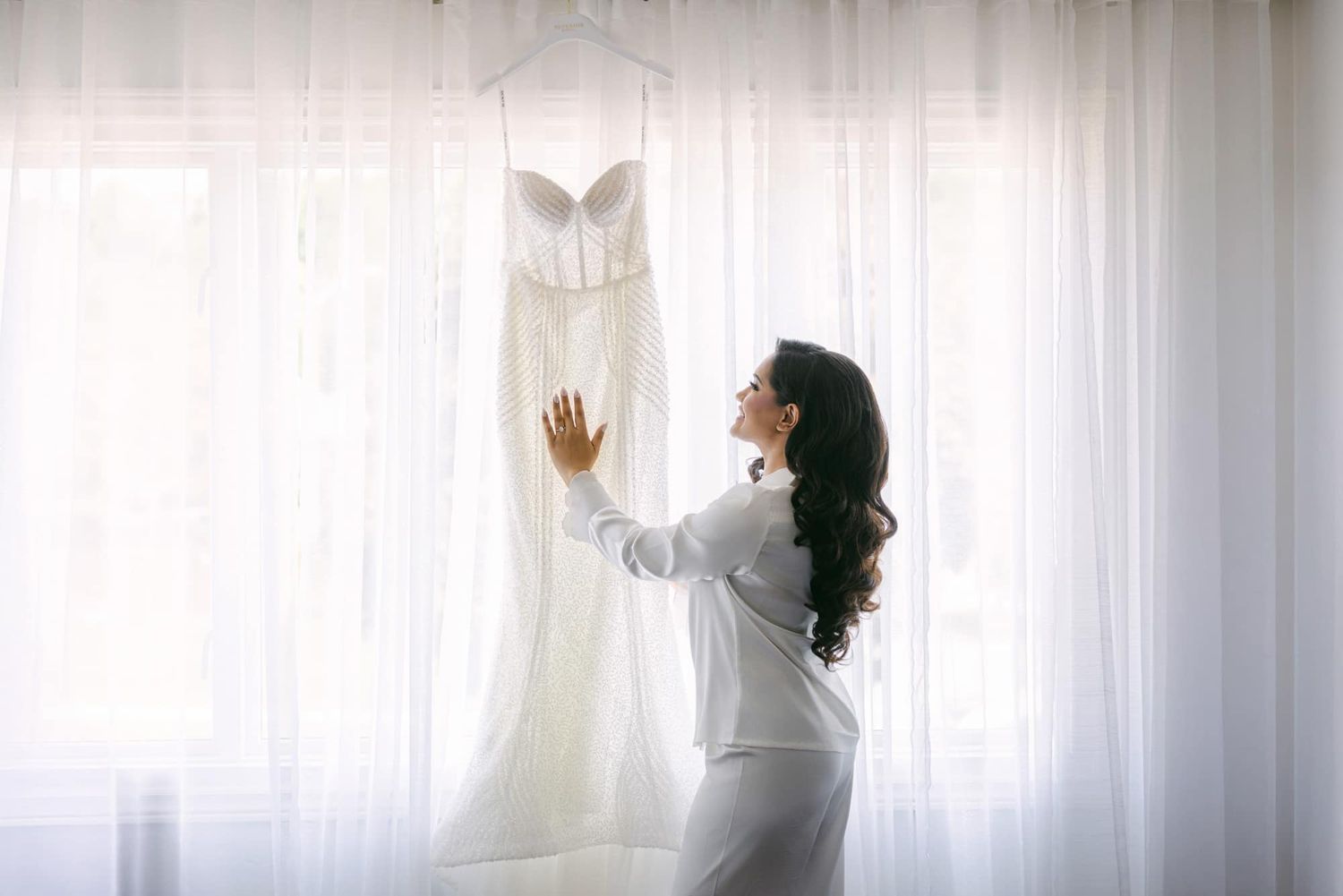 Bride in white robe admires her strapless wedding gown hanging by a sunlit window before getting dressed.