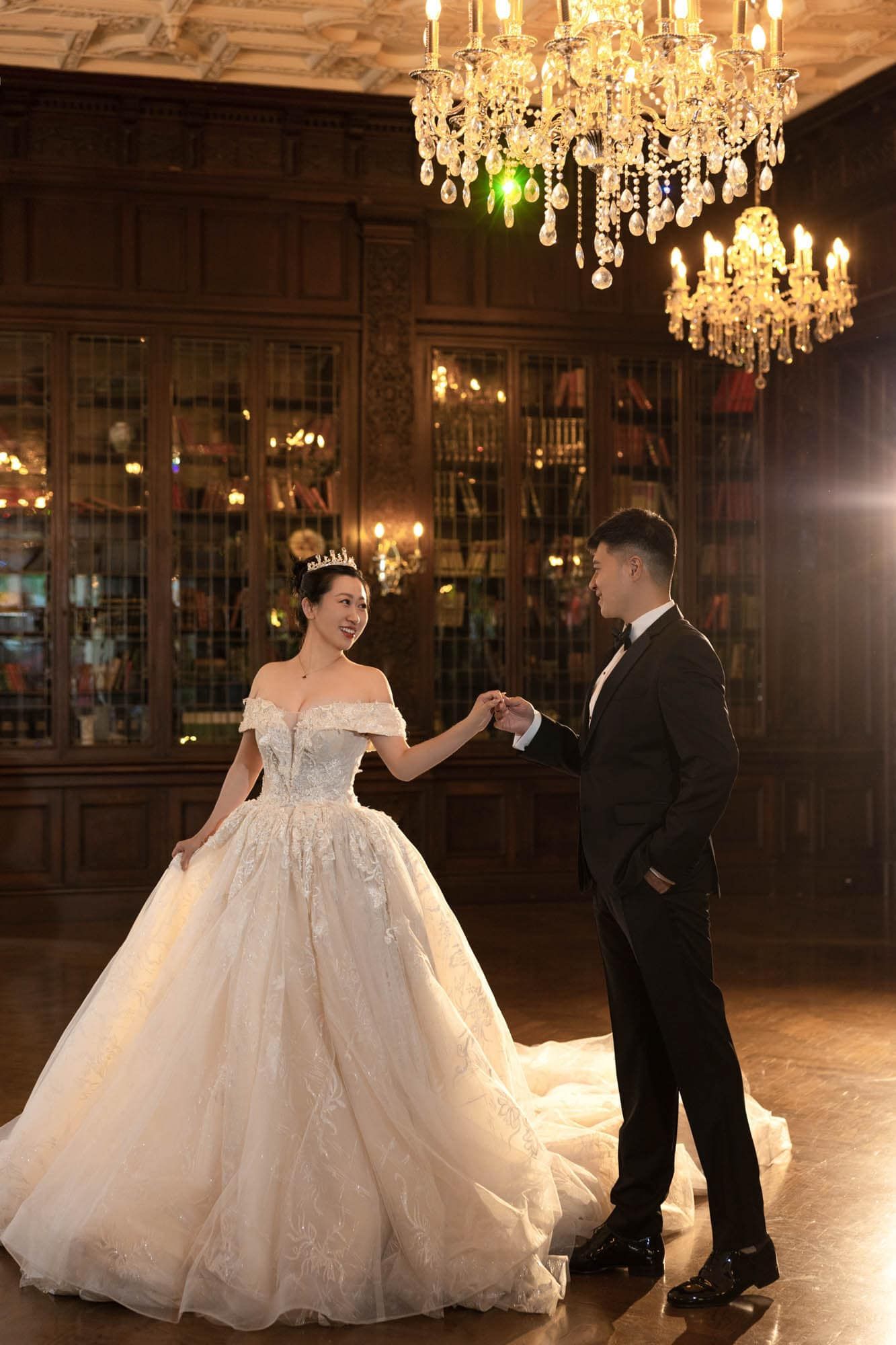 Bride in off-shoulder ballgown and groom dancing beneath a golden chandelier in an elegant library.