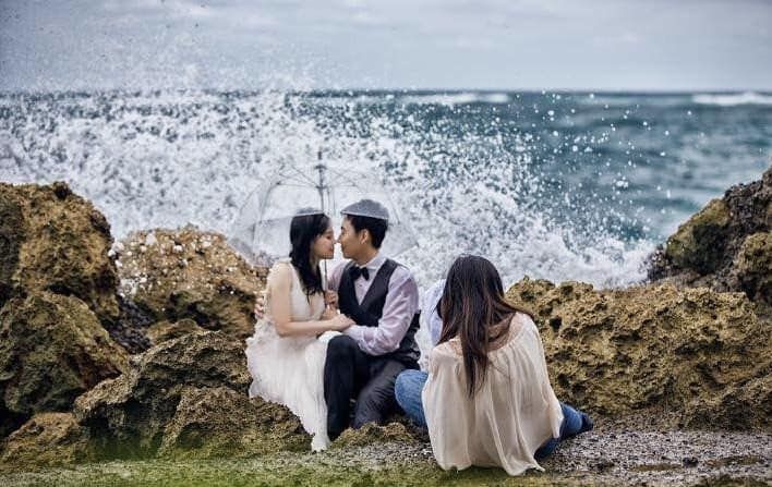 Bride and groom kissing on rocky shore with crashing ocean waves behind them.