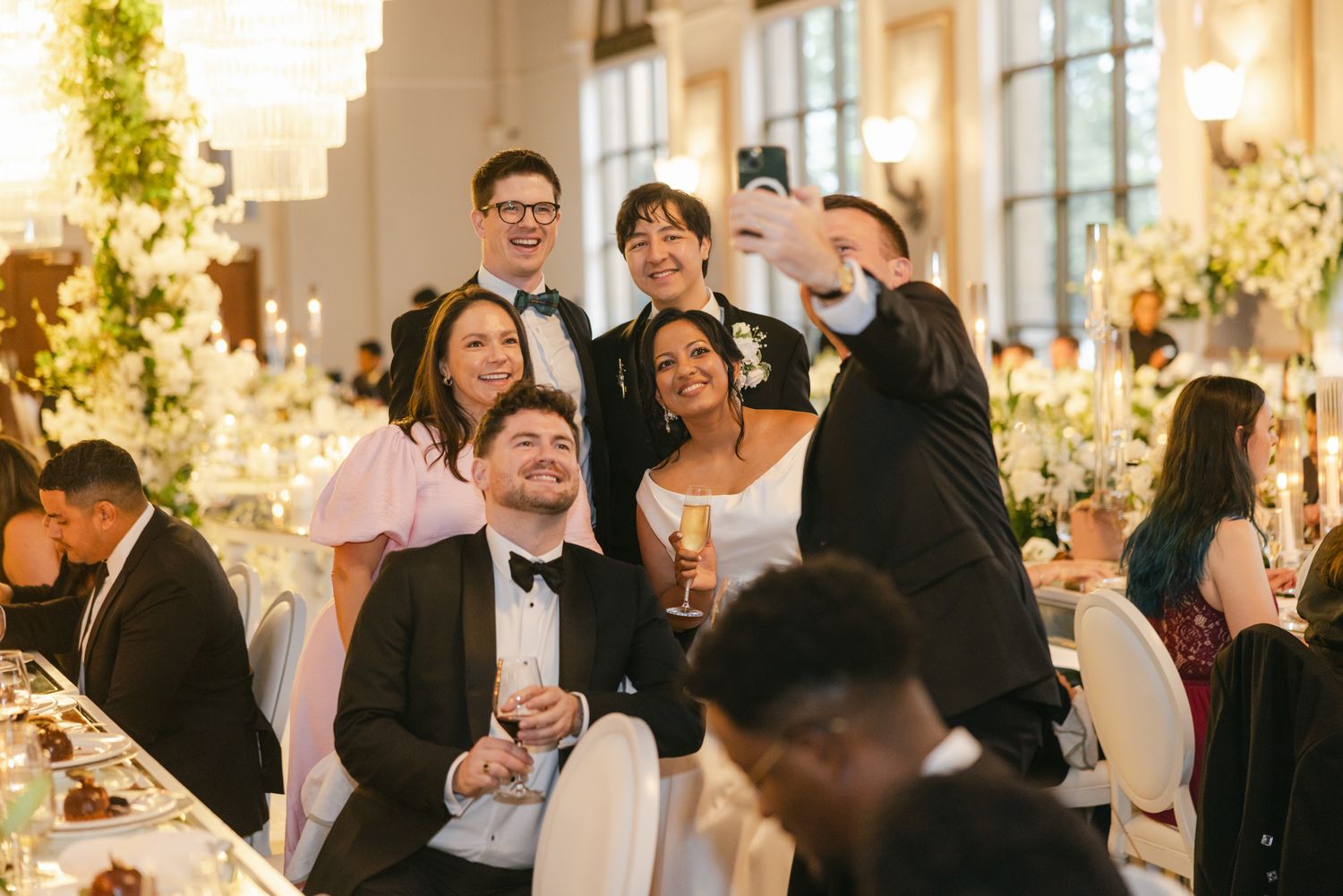 Wedding guests in formal attire pose for a group selfie at a reception table with flowers and a candlelit ballroom.